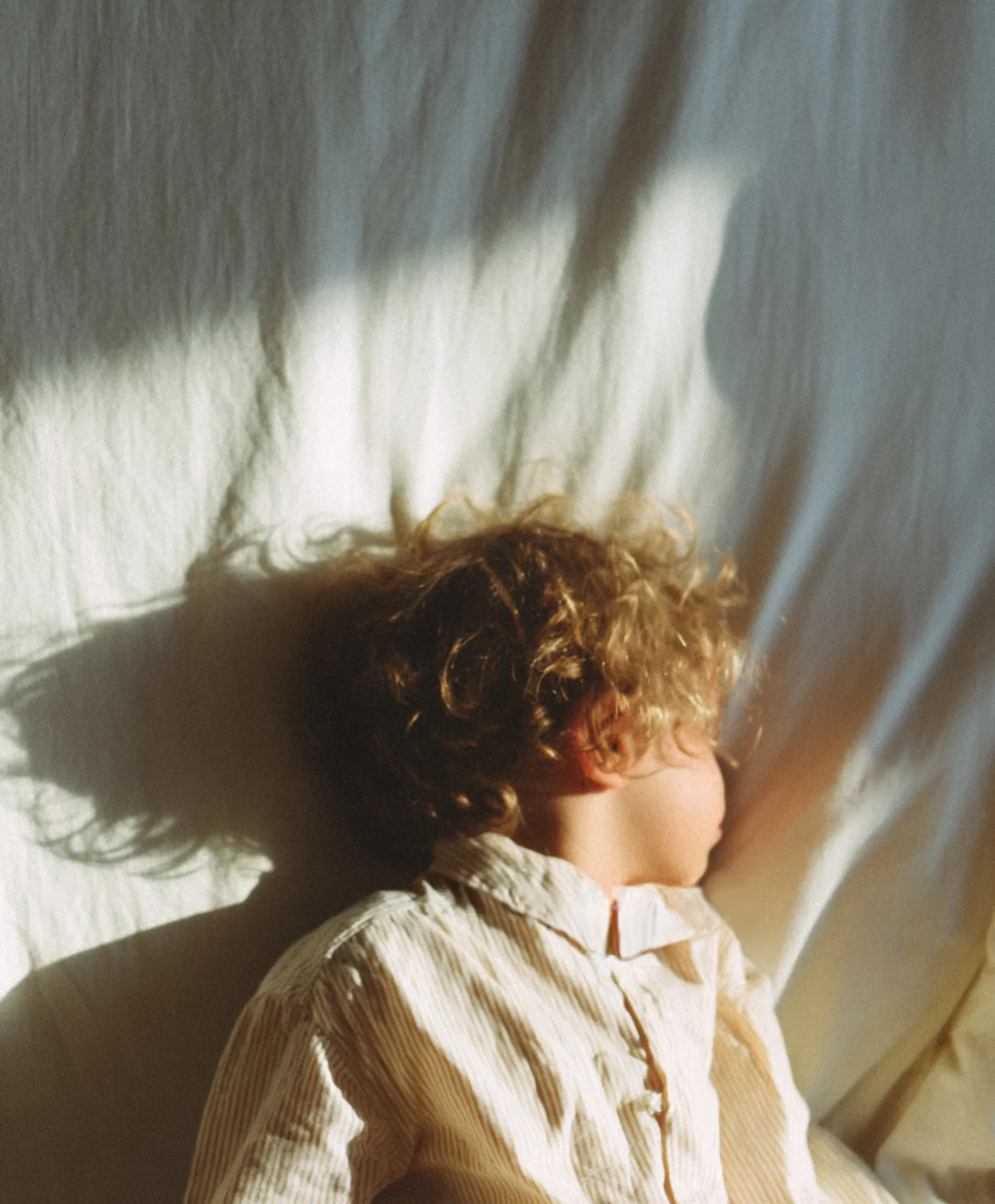 A child in a striped nightshirt sleeps in morning light, the shadow of curly hair across the sheet.