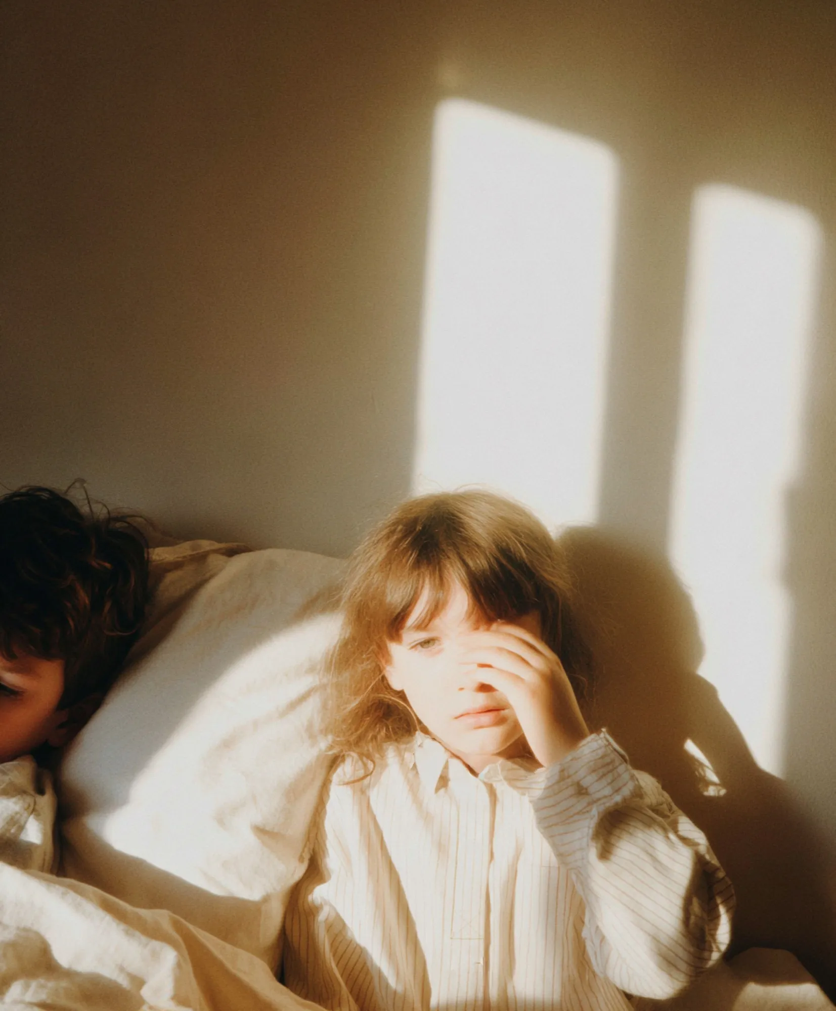 A child in a striped nightshirt sleeps in morning sun, the shadow of curly hair cast across the crumpled sheet.